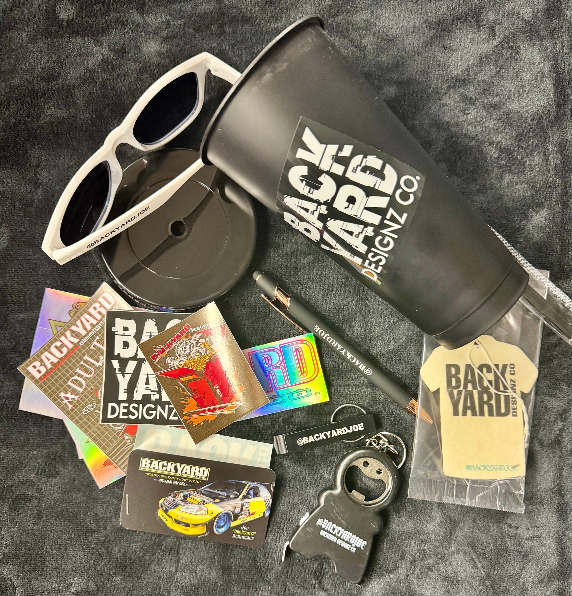 Collection of Backyard Designz Co. branded items including sunglasses, tumbler, keychain, and more on a dark textured background.